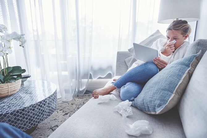 Woman sitting on the sofa with a cold, flu or allergy. She is using her tablet to do a Google search for a healthcare provider in her area. There are tissues on the sofa around her.