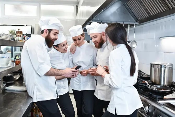 Cooking team in restaurant kitchen discussing something while holding their mobile smartphones.